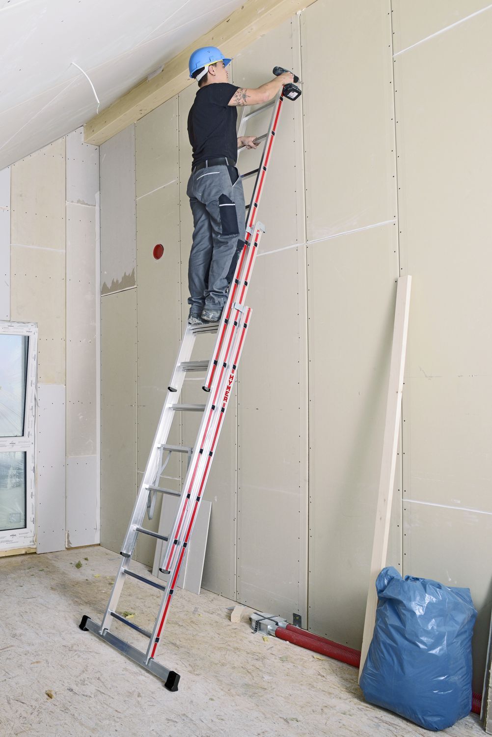 Handwerker auf einer dreiteiligen Mehrzweckleiter von Hymer stehend bei Arbeiten an einer Wand in einem Rohbau