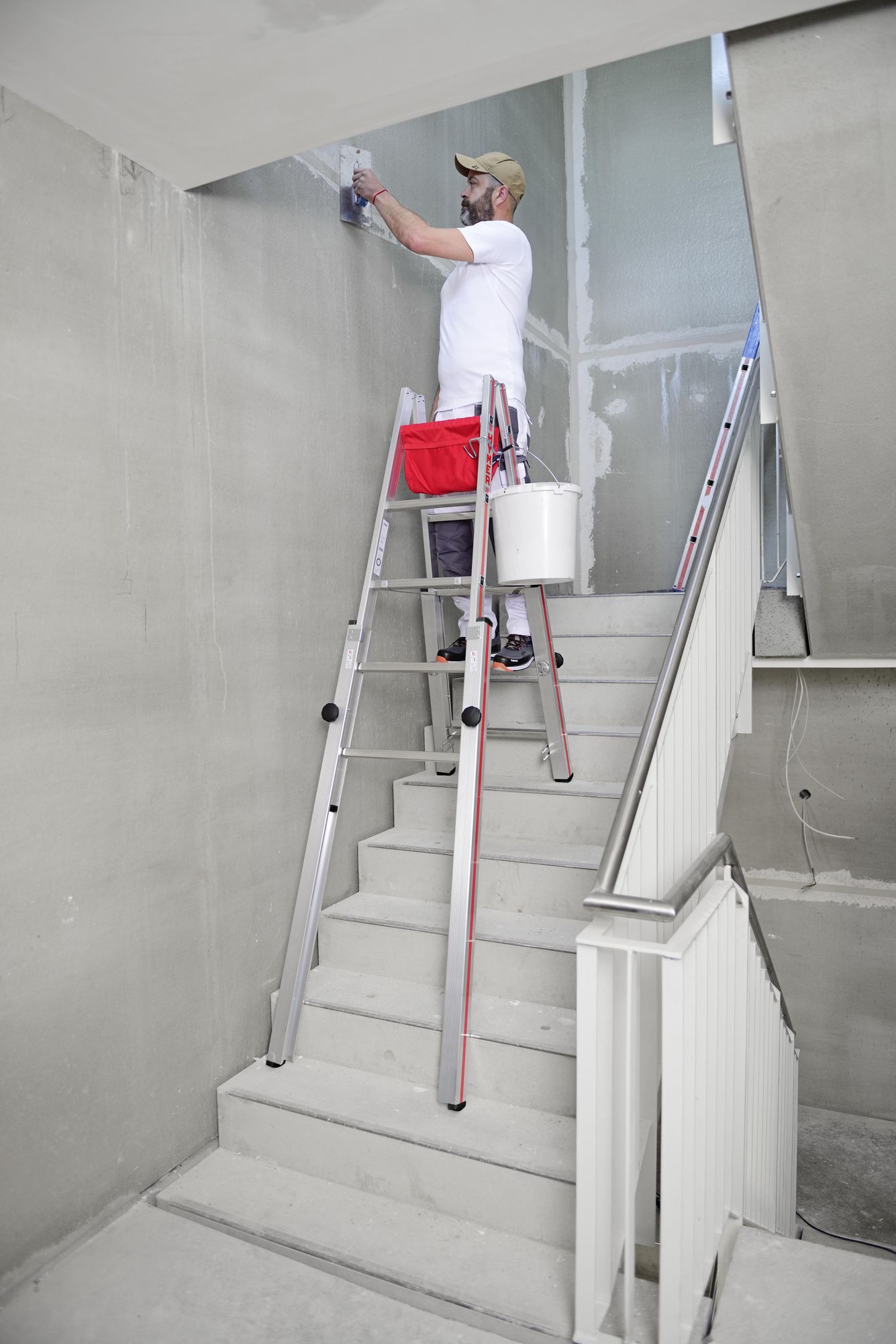 Hymer Treppenstehleiter 2x 5 Sprossen Modell 412310 Anwendungsbeispiel Maler steht bei Arbeiten in einem Rohbau-Treppenhaus auf einer Hymer Treppenstehleiter 2x 5 Sprossen Modell 412310, die auf einer Treppe steht bild 2
