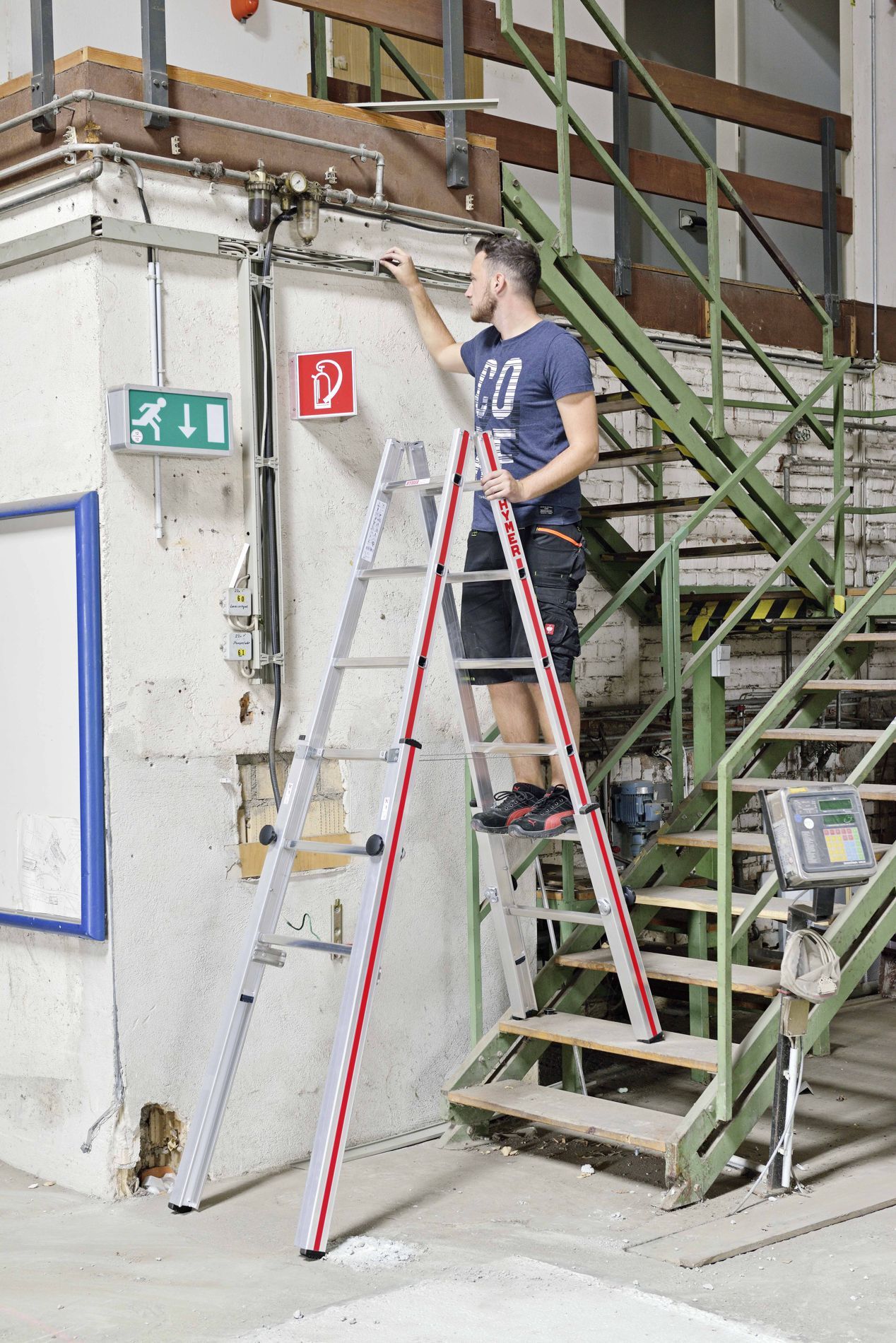 Hymer Treppenstehleiter 2x 6 Sprossen Modell 412312 Anwendungsbeispiel Handwerker in Arbeitskleidung auf einer Hymer Treppenstehleiter stehend bei Montagearbeiten in einer Fabrikhalle