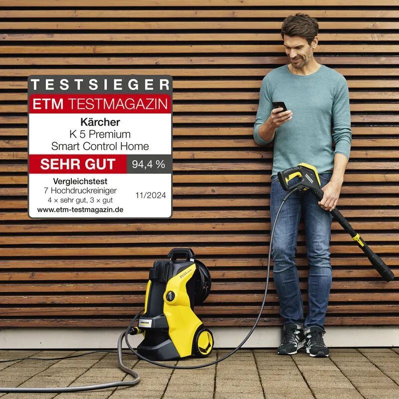 Kärcher Hochdruckreiniger K 5 Premium Smart Control Home Auszeichnung ETM Testmagazin 11/2024 Ein Mann lehnt an einer holzvertäfelten Wand im Freien und schaut auf sein Handy, in der Hand hat er die Pistole des Kärcher Hochdruckreinigers, der neben ihm auf dem gepflasterten Boden steht