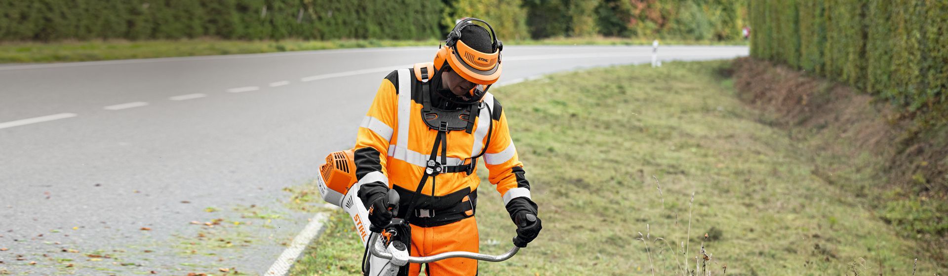Mann in orangefarbener Stihl Warnschutzkleidung mit Schutzhelm bei Mäharbeiten an einer Landstraße
