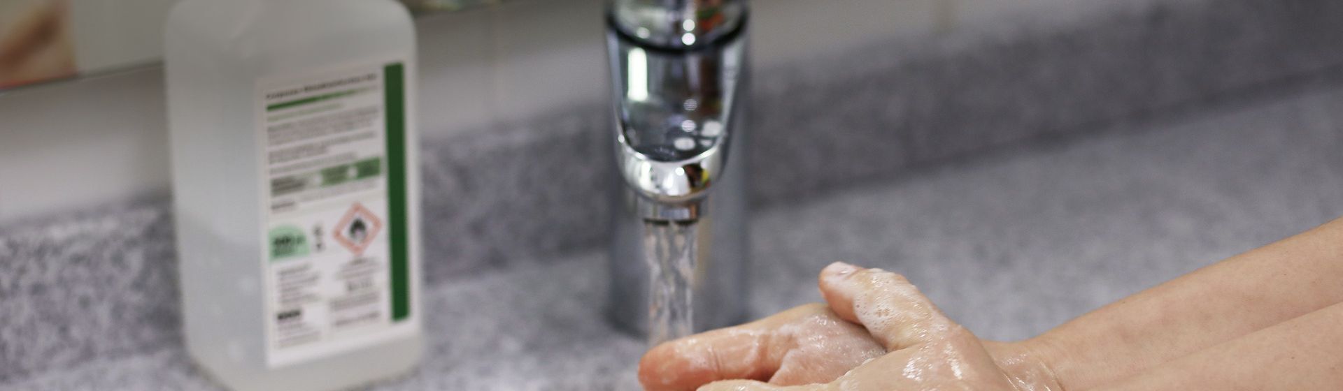 Händewaschende Person an einem Waschbecken, im Hintergrund ein Desinfetionsmittelspender auf dem Rand stehend Händewaschende Person an einem Waschbecken, im Hintergrund ein Desinfetionsmittelspender auf dem Rand stehend