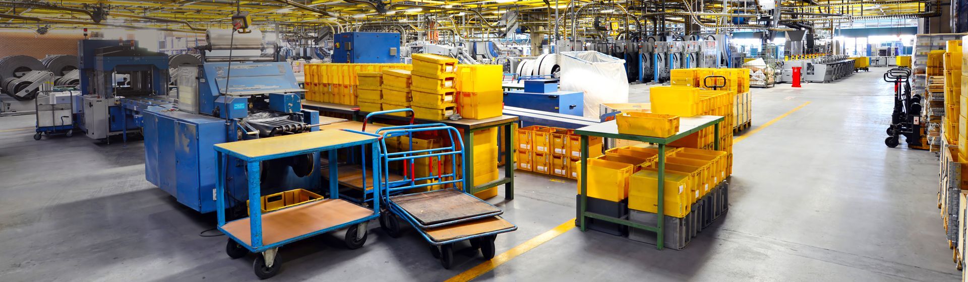 Blick in die Produktion eines Unternehmens, zu sehen sind Maschinen, gelbe Postbehälter, Transportwagen und Hubwagen Blick in die Produktion eines Unternehmens, zu sehen sind Maschinen, gelbe Postbehälter, Transportwagen und Hubwagen