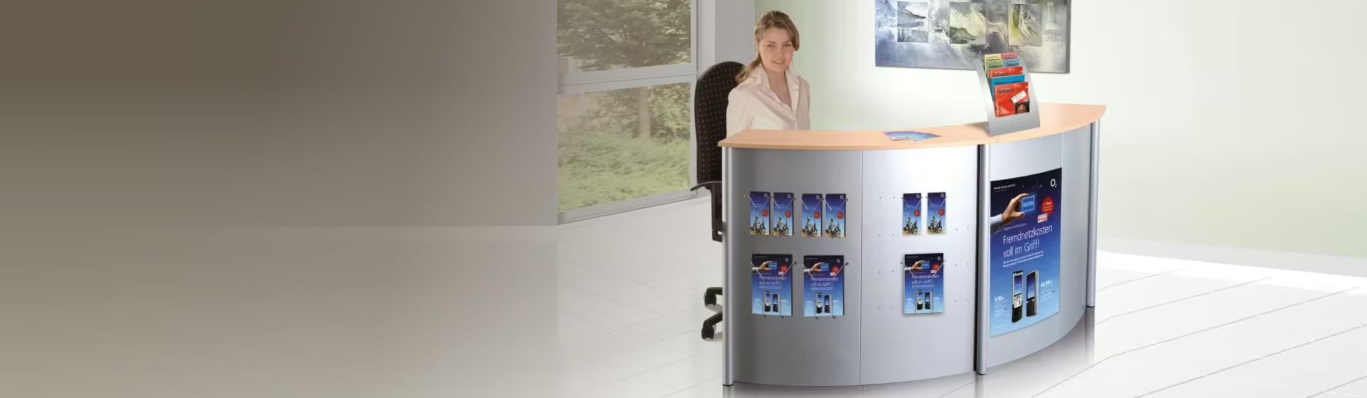 Eine Frau an einem halbrunden Empfangs-Thekensystem mit silberfarbenen Metallelemente und Holztheke sitzend, auf dessen Front Werbeplakate und Broschürenhalterungen angebracht sind Eine Frau an einem halbrunden Empfangs-Thekensystem mit silberfarbenen Metallelemente und Holztheke sitzend, auf dessen Front Werbeplakate und Broschürenhalterungen angebracht sind