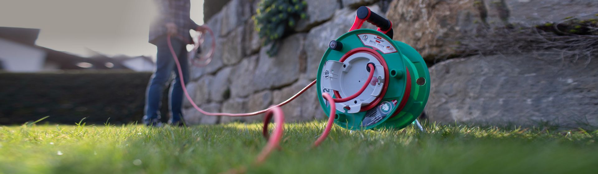 Grüne Brennenstuhl Garten-Kabeltrommel mit ausgerolltem roten Kabel stehend auf einer Rasenfläche vor einer Mauer Grüne Brennenstuhl Garten-Kabeltrommel mit ausgerolltem roten Kabel stehend auf einer Rasenfläche vor einer Mauer