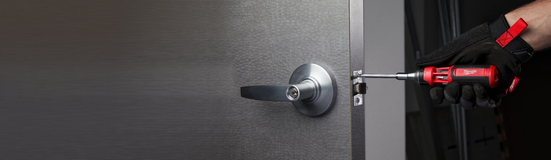 Handwerker befestigt einen Schliesser an der Seite einer grauen Tür mit einem Wechselklingen Schraubendreher von Milwaukee Handwerker befestigt einen Schliesser an der Seite einer grauen Tür mit einem Wechselklingen Schraubendreher von Milwaukee