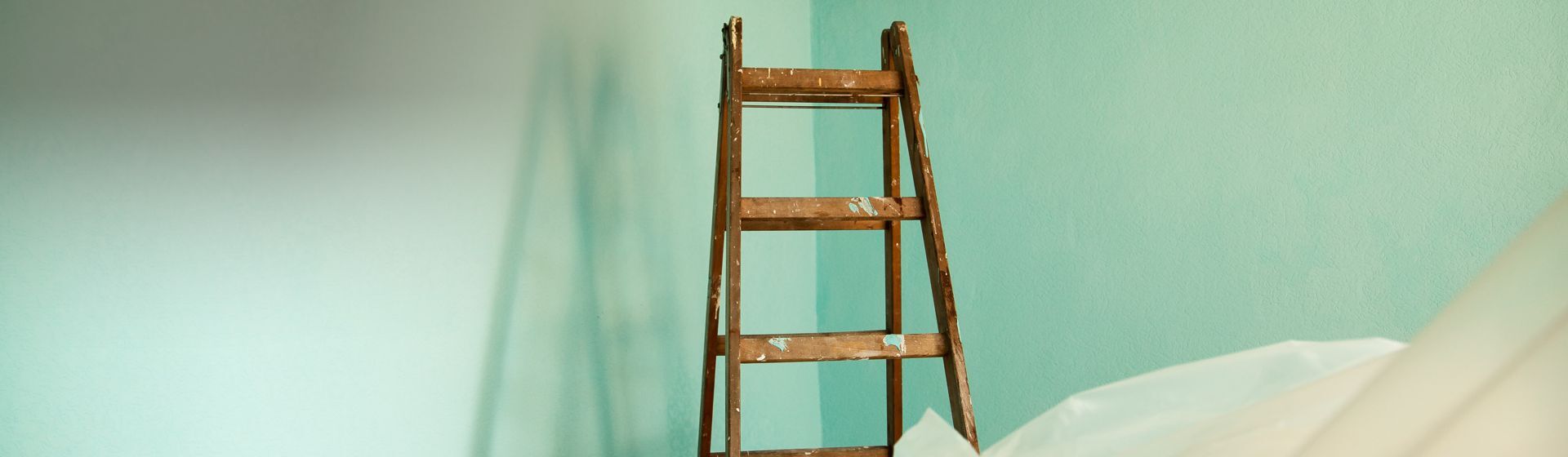 Farbverschmierte braune Holzleiter in einem renovierungsbedürftigen Raum mit einer mintgrünen Wand Farbverschmierte braune Holzleiter in einem renovierungsbedürftigen Raum mit einer mintgrünen Wand